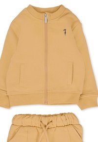 Giacca zip-up marrone chiaro con due tasche laterali e un piccolo logo. Abbinata a pantaloni con elastico in vita dello stesso marrone chiaro. Tessuto morbido.