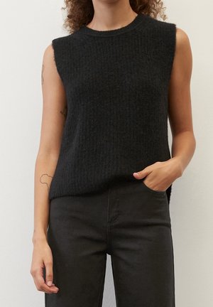 Woman wearing a sleeveless black textured knit top and black pants, with hand in pocket, standing against a plain light background.