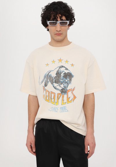 Beige oversized t-skjorte med grafikk av en brølende panter, oransje og blå tekst, og fem gule stjerner. Brukt med svarte bukser.