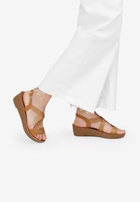 Sandali con zeppa marroni e cinturini incrociati, con una finitura testurizzata e una fibbia sicura alla caviglia. Mostrati su un piede, abbinati a pantaloni bianchi a gamba larga.