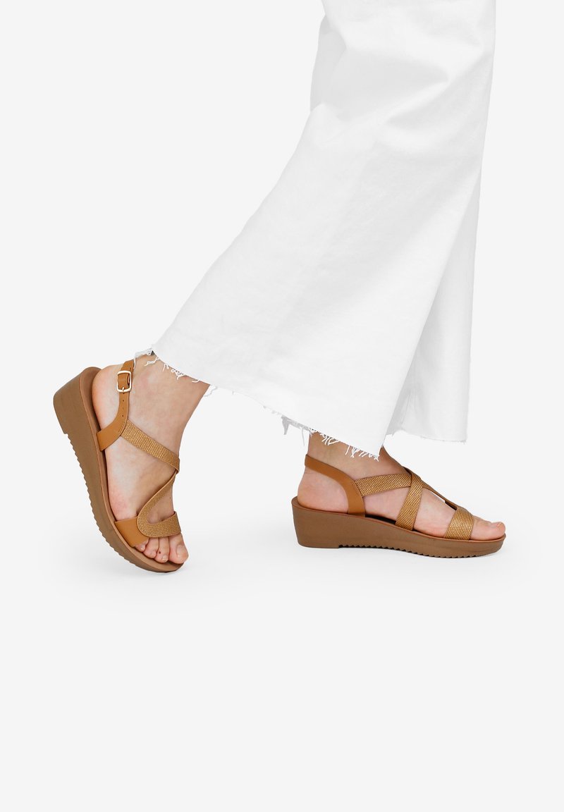 Sandali con zeppa marroni e cinturini incrociati, con una finitura testurizzata e una fibbia sicura alla caviglia. Mostrati su un piede, abbinati a pantaloni bianchi a gamba larga.