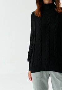 Pull en tricot noir avec un col montant, des manches longues et un ourlet à côtes ; associé à un pantalon tailleur gris clair, avec une finition texturée.