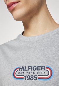 Camiseta de algodón gris con un logo impreso multicolor: "HILFIGER NEW YORK CITY 1985" con acentos en azul, rojo y blanco. Diseño de cuello redondo.