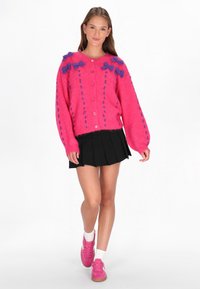 Cardigan tricotat roz cu accente de flori mov, având guler rotund și închidere cu nasturi. Asortat cu o fustă plisată neagră și adidași roz.