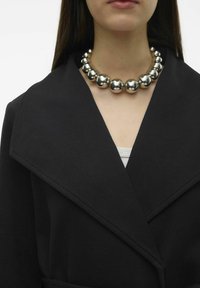 Silverfärgad sfärisk halsband med glänsande yta, bärs över en svart kappa med bred krage och strukturtrikå, som framhäver dess design.