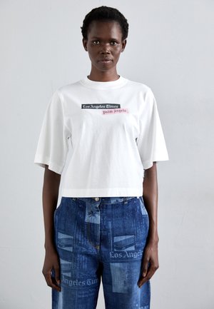 Camiseta corta blanca con el texto "Los Angeles Times" y "Palm Angels" en negro y rosa. Combinada con unos vaqueros azules con patrones de impresión de periódico.