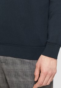 Donkerblauwe geribbelde mouw met een getailleerd ontwerp, gecombineerd met grijs geruite broeken. De texturen zijn glad en gestructureerd.