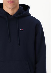 Temno modra kapuca iz mehkega blaga. Ima kenguru žep, vrvico za zategovanje kapuce in majhen vezen logotip v rdeči, beli in modri barvi.