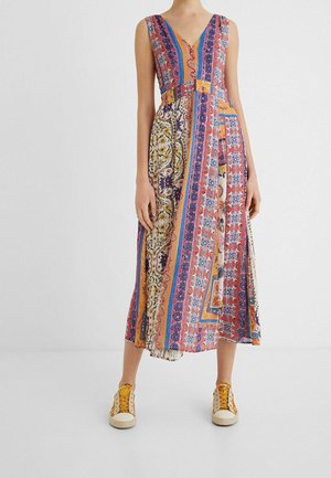 Vestido midi sin mangas con patrones coloridos de patchwork combinado con zapatillas blancas y amarillas en una persona de pie.