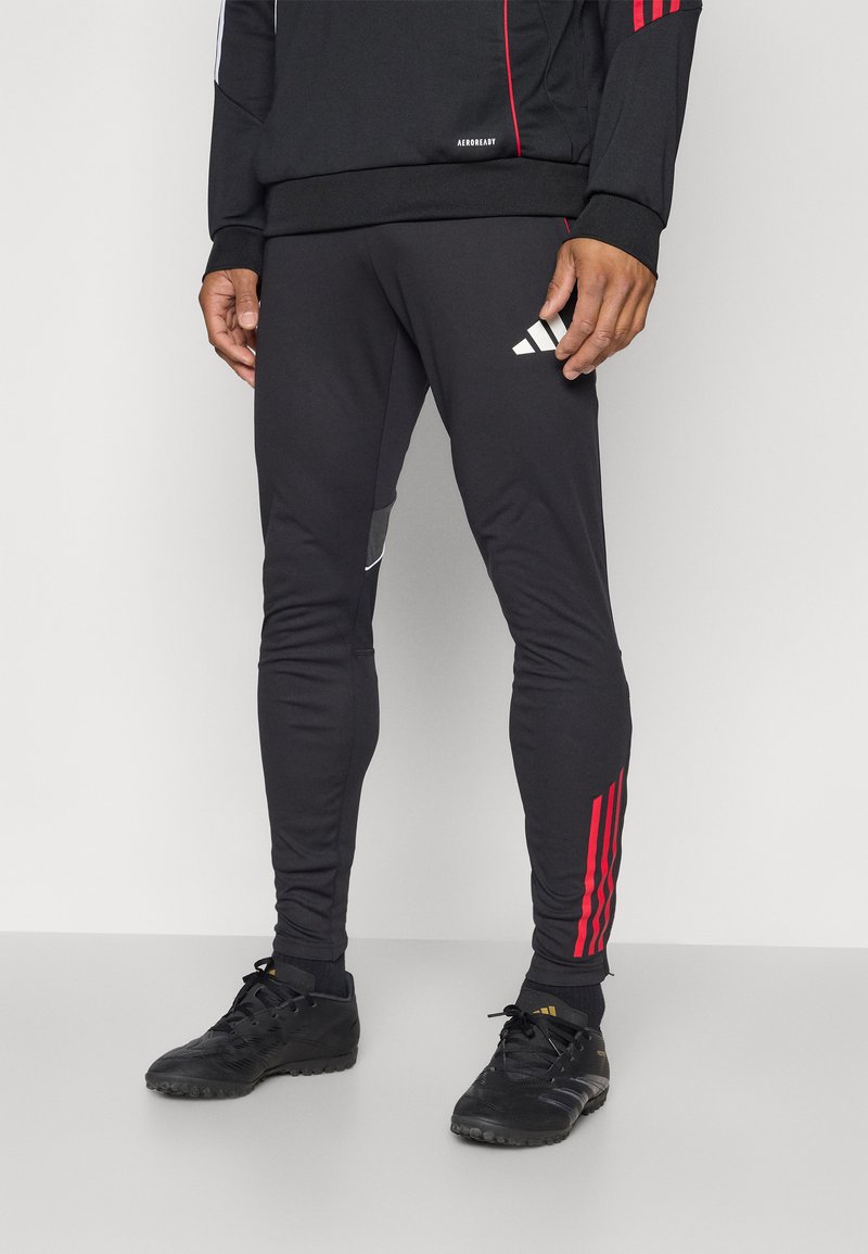 Pantalones deportivos negros de corte ajustado, con franjas rojas y blancas, y un logo negro en el muslo izquierdo. Hechos de tejido elástico.