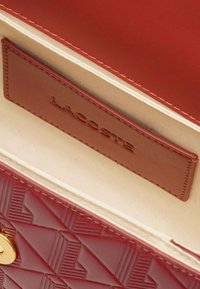 Intérieur d'un sac à main en cuir rouge montrant un motif gaufré, des accessoires en or et une fente pour carte marron portant l'inscription "LACOSTE". Doublure beige clair visible.