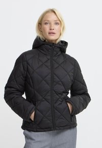 Femme blonde portant une veste matelassée noire avec capuche, les mains dans les poches zippées, et un pantalon gris clair sur un fond blanc.