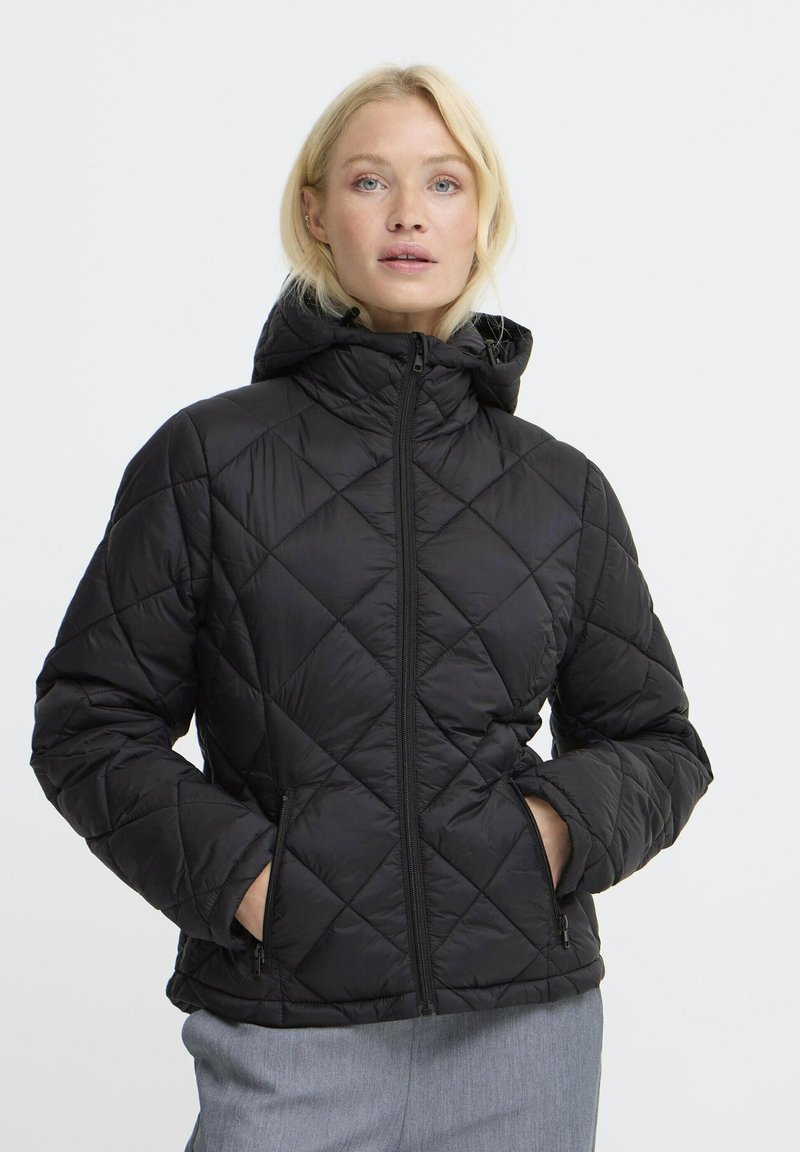 Femme blonde portant une veste matelassée noire avec capuche, les mains dans les poches zippées, et un pantalon gris clair sur un fond blanc.