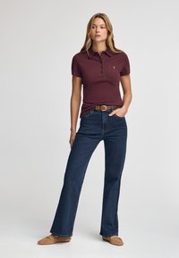 Maroonowy polo z krótkim rękawem z kołnierzykiem i trzema guzikami, w zestawie z ciemnoniebieskimi dżinsami dzwonami i beżowymi mokasynami. Gładka tekstura tkaniny.