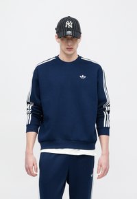 Μπλε σκούρο φούτερ με λευκό λογότυπο Adidas, με λευκές ρίγες κατά μήκος των μανικιών. Συνδυάζεται με παντελόνι σετ και καπέλο με μαύρες ρίγες.