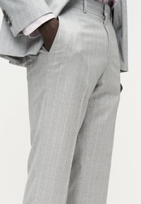 Άνδρας που φοράει παντελόνι με ανοιχτό γκρι λεπτές ρίγες, με τα χέρια στις τσέπες, εμφανιζόμενος από τη μέση έως τη μέση μηρών πάνω σε απλό φόντο.