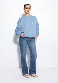Jasnoniebieska bluza z okrągłym dekoltem i małym logo na piersi, w połączeniu z rozkloszowanymi niebieskimi dżinsami i jasnoróżowymi butami, na białym tle.