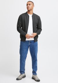 Chaqueta bomber gris oscuro con textura acanalada, cierre de cremallera y acentos negros, llevada sobre una camiseta blanca, combinada con jeans azules y zapatos grises.