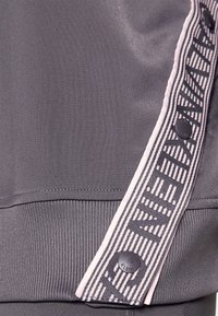 Gray athletic top featuring a textured fabric with a striped ribbon along the side, accented by a branded black button.