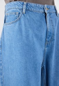 Calças de ganga de alta cintura em azul claro com um corte descontraído, apresentando um bolso frontal, costuras amarelas e fecho de botão.