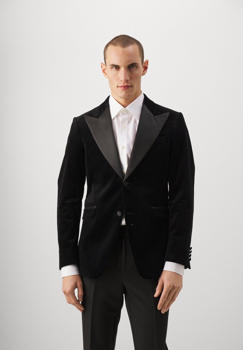 Czarna aksamitna marynarka smokingowa z satynowymi klapami w kształcie szala, z pojedynczym guzikiem, dwoma przednimi kieszeniami i teksturowanym wykończeniem. Biała koszula widoczna pod spodem.