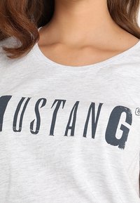 Lysegrå T-shirt med blød tekstur, prydet med fed mørk tryk af "MUSTANG" i varierende skrifttyper henover brystet. Rund halsudskæring.