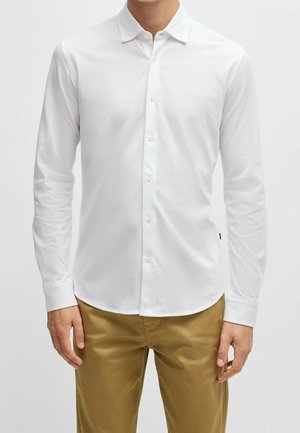 Mann trägt ein eng anliegendes weißes langärmeliges Hemd und hellbraune Hose vor einfachem Hintergrund.