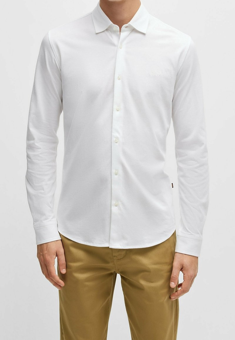 Homme portant une chemise blanche cintrée à manches longues boutonnée et un pantalon marron clair sur un fond uni.