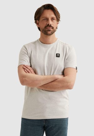 Man met korte baard en middengrijsbruin haar, draagt een lichtgrijs T-shirt met korte mouwen en blauwe spijkerbroek, staand met gekruiste armen.