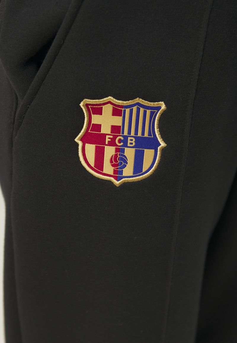 Pantaloni neri con un ricamo del crest del FC Barcelona in rosso, blu e oro, che presenta una forma a scudo con strisce orizzontali e verticali.