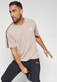 Lys beige, kortermet treningstrøye med svarte sidestiper og hvit logo, kombinert med mørke bukser og holder en kettlebell.