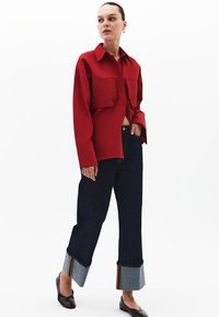 Camicia rossa con due tasche frontali, abbinata a pantaloni jeans larghi scuri con orli grigi e dettagli a righe. Ballerine nere completano l'outfit.
