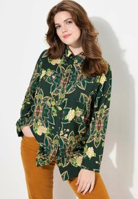 Green floral-patterned button-up shirt featuring large blossoms and intricate designs, paired with mustard-colored pants. Soft fabric texture.