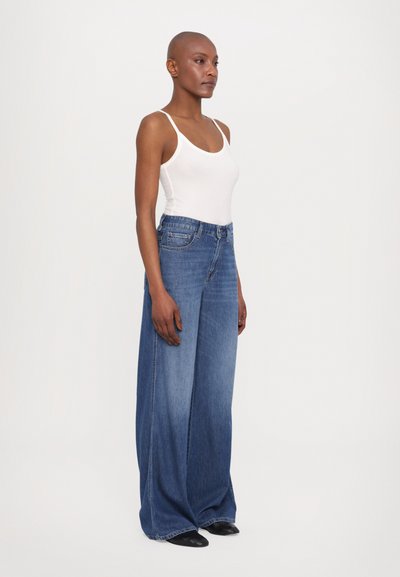 Wide-leg denim jeans in medium blue, with a high waist, polished finish, and front pockets. Paired with a fitted white tank top.