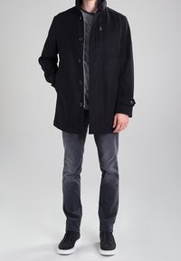 Cappotto nero, con bottoni e colletto alto, realizzato in un tessuto liscio. Indossato sopra una camicia grigia, abbinato a pantaloni grigi e sneakers nere.