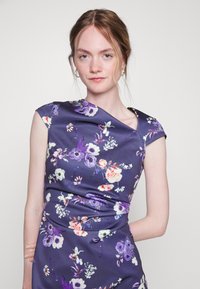 Vestido floral em satin púrpura com um design ajustado, mangas compridas e detalhes franzidos. Apresenta vários padrões florais coloridos.