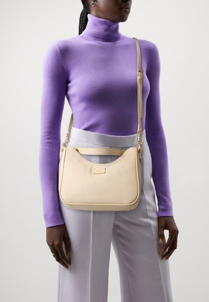 Beige Umhängetasche mit goldenen Beschlägen, die ein geschwungenes Design und eine strukturierte Oberfläche aufweist. Das Modell trägt einen lila Rollkragenpullover und helle Hosen.