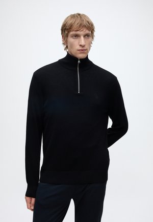 Blond man wearing a black long-sleeve pullover with a half-zip collar, standing against a plain light background with one hand behind his back.