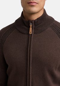 Brązowy sweter dzianinowy z wysokim, prążkowanym kołnierzem, zapięciem na zamek oraz teksturowanymi akcentami na ramionach. Posiada gładką tkaninę i dopasowany krój.