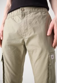 Světlé khaki cargo kalhoty s elastickým pasem, bočními kapsami a malou nášivkou na stehně. Texturovaná, odolná tkanina.