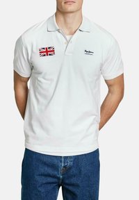 Biela polo košeľa s preloženým golierom, s nášivkou Union Jack na ľavej hrudi a vyšitým logom značky v čiernej farbe. Klasický strih.