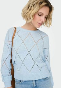 Pull en tricot bleu clair avec un motif en diamant et des détails à points ajourés, manches avec bordures côtelées, porté avec un jean en denim.