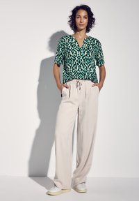 Camicia a maniche corte verde con motivo a diamante e foglie, abbinata a pantaloni a gamba larga beige chiaro e sneakers bianche.