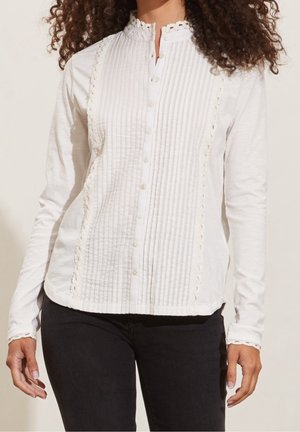 Mujer que lleva una blusa blanca de manga larga con plisados verticales, adornos de encaje y botones en la parte delantera, combinada con pantalones negros.