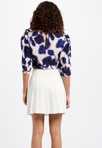 Top met opgeblazen mouwen met een paarse en blauwe bloemenprint, gecombineerd met een witte geplooide rok, gezien vanaf de achterkant.