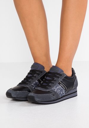 Nogi w czarno-szarych sneakersach Versace Jeans z błyszczącymi akcentami i czarnymi sznurówkami na białej podłodze.