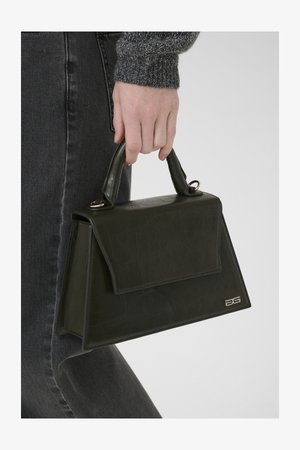 Mão segurando uma pequena bolsa verde-escura com uma superfície texturizada e um logótipo metálico DG, usada com jeans pretos e uma manga de malha cinza.