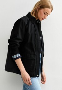 Svart jacka av bomull med ett kordflöjelskrage, dragkedja framtill och sidofickor. Har uppkavlade ärmar med randiga manchetter.