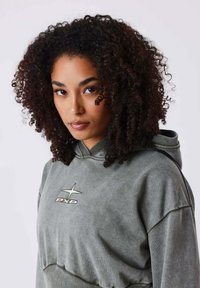 Lichtgroene hoodie met een logo aan de voorkant, trekkoordkap en zachte textuur. De mouwen zijn wijd met een ontspannen pasvorm.