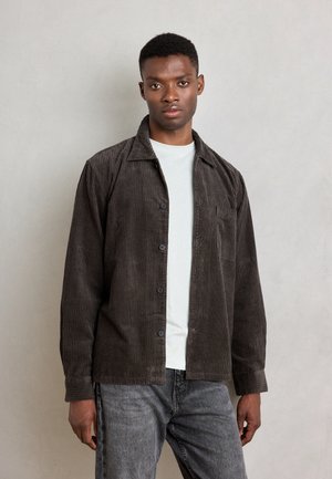 Corduroy shirt in dark brown with button-up front, pointed collar, and chest pocket. Layered over a light grey t-shirt, paired with grey denim.